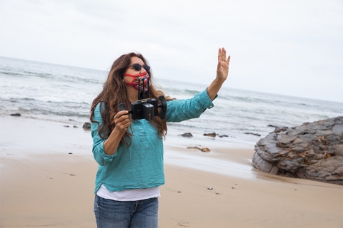 Image of woman on beach with mask/face covering, video camera in hand, arm up as if directing an act
